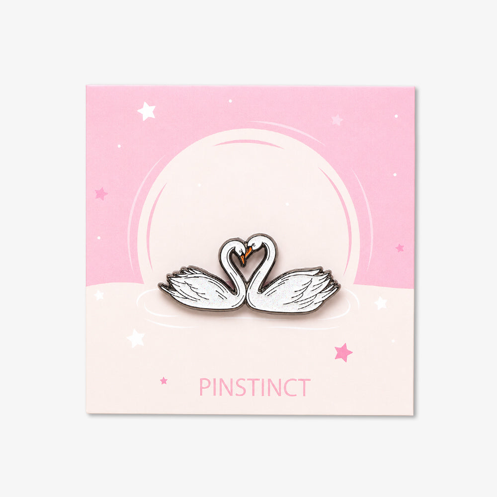 Enamel pin featuring two white swans with orange beaks forming a heart shape, mounted on a pink and cream backing card with star details