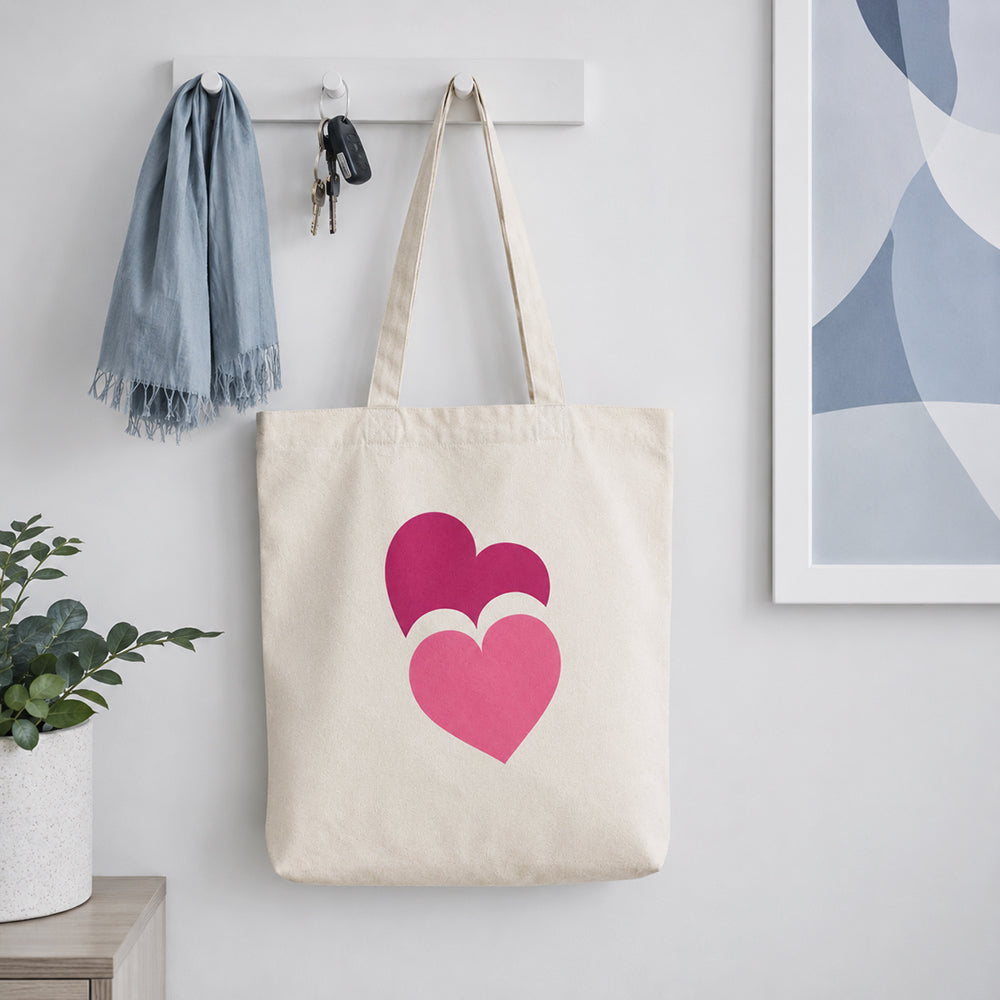 Basic canvas tote bag in natural beige with two overlapping pink heart prints, hanging on a wall hook at home