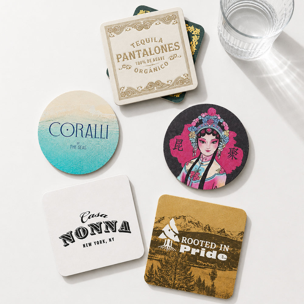 Custom pulpboard coasters with diverse custom printed designs, round and square shapes, arranged on white table beside a clear water glass