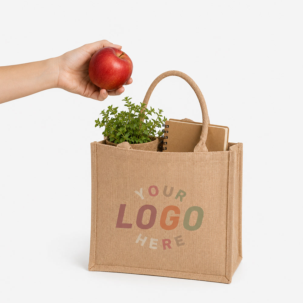 Natural jute tote bag with custom logo placeholder, holding potted plant and notebook beside a hand holding red apple