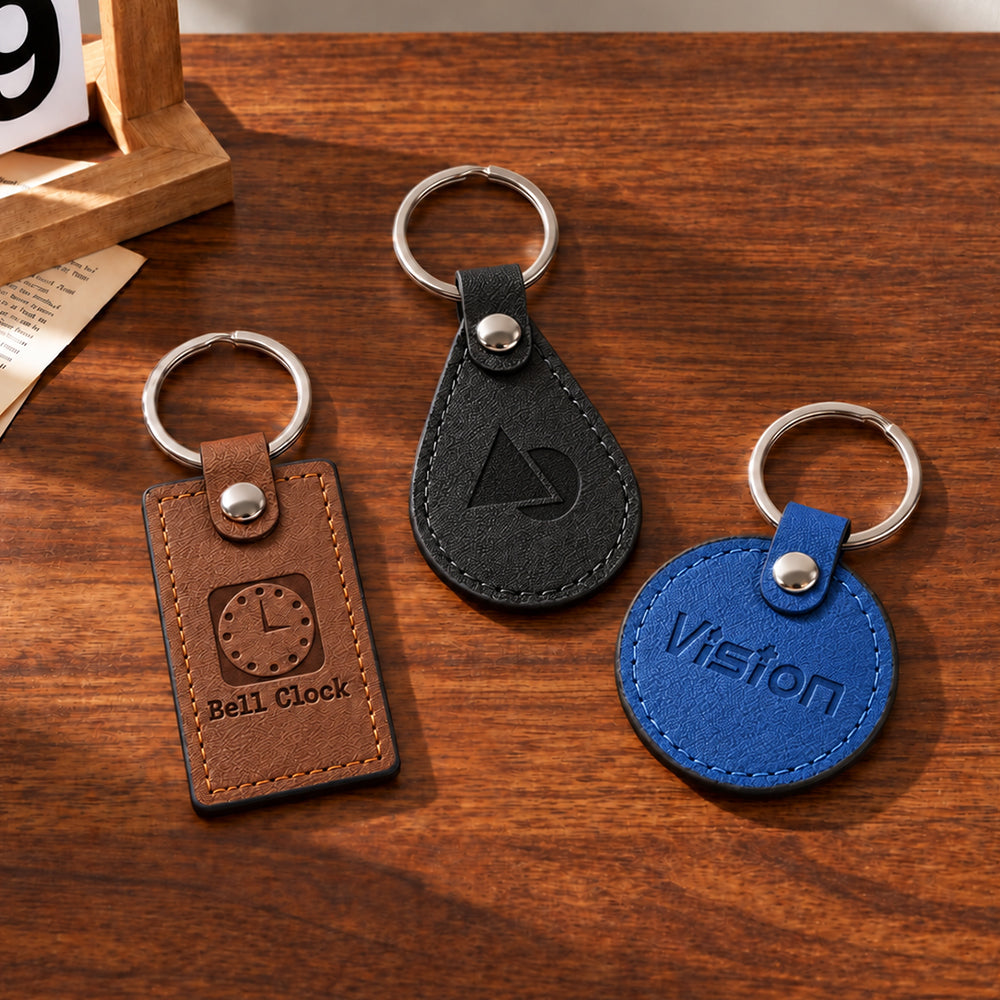 Three PU leather keychains in brown rectangle, black teardrop, and blue circle shapes with stitched edges and metal rings
