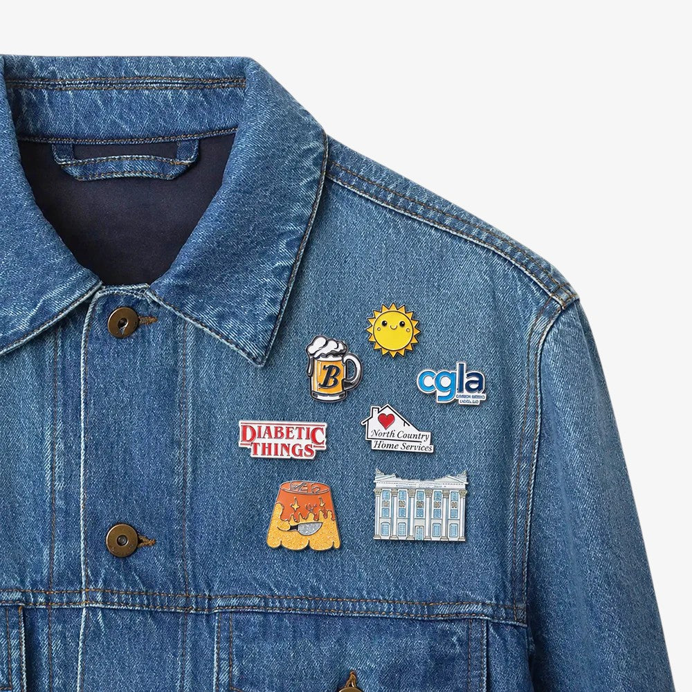 Soft enamel pins on denim: a smiling sun, beer mug, flame-topped dessert, house with heart, and classical building, all with colorful fills and metal outlines