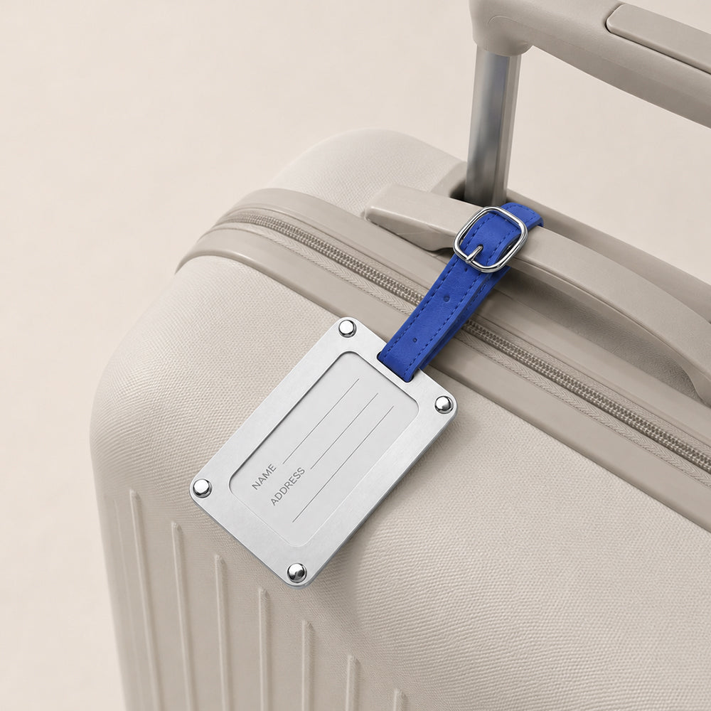 Stainless Steel luggage tag with blue leather strap, attached to beige suitcase, with reserved name and address writing area